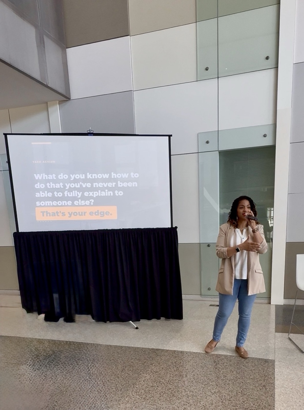 Java Hardge speaking with a microphone in front of a presentation slide reading 'Take action: what do you know how to do that you've never been able to fully explain to someone else? That's your edge.'
